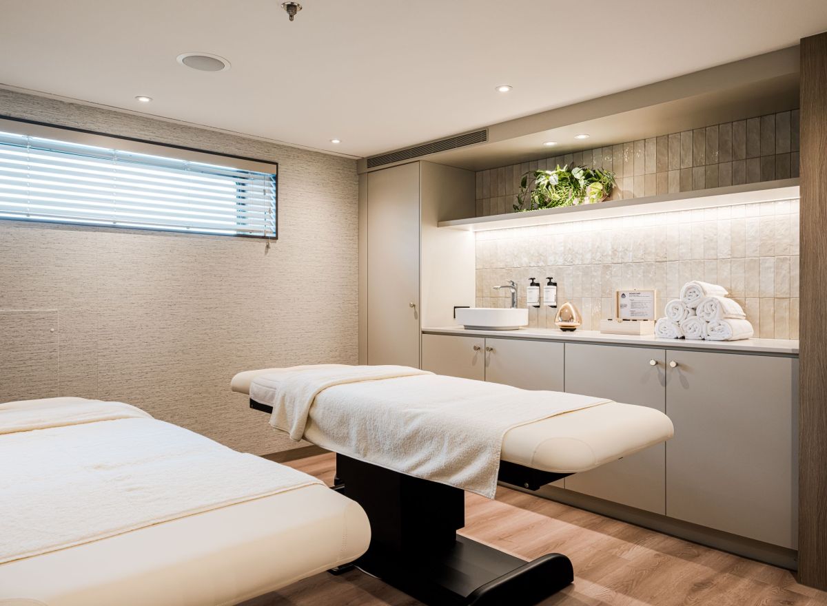 Calm and elegant wellness room with two treatment beds, soft lighting, rolled towels, and a modern counter with spa amenities.