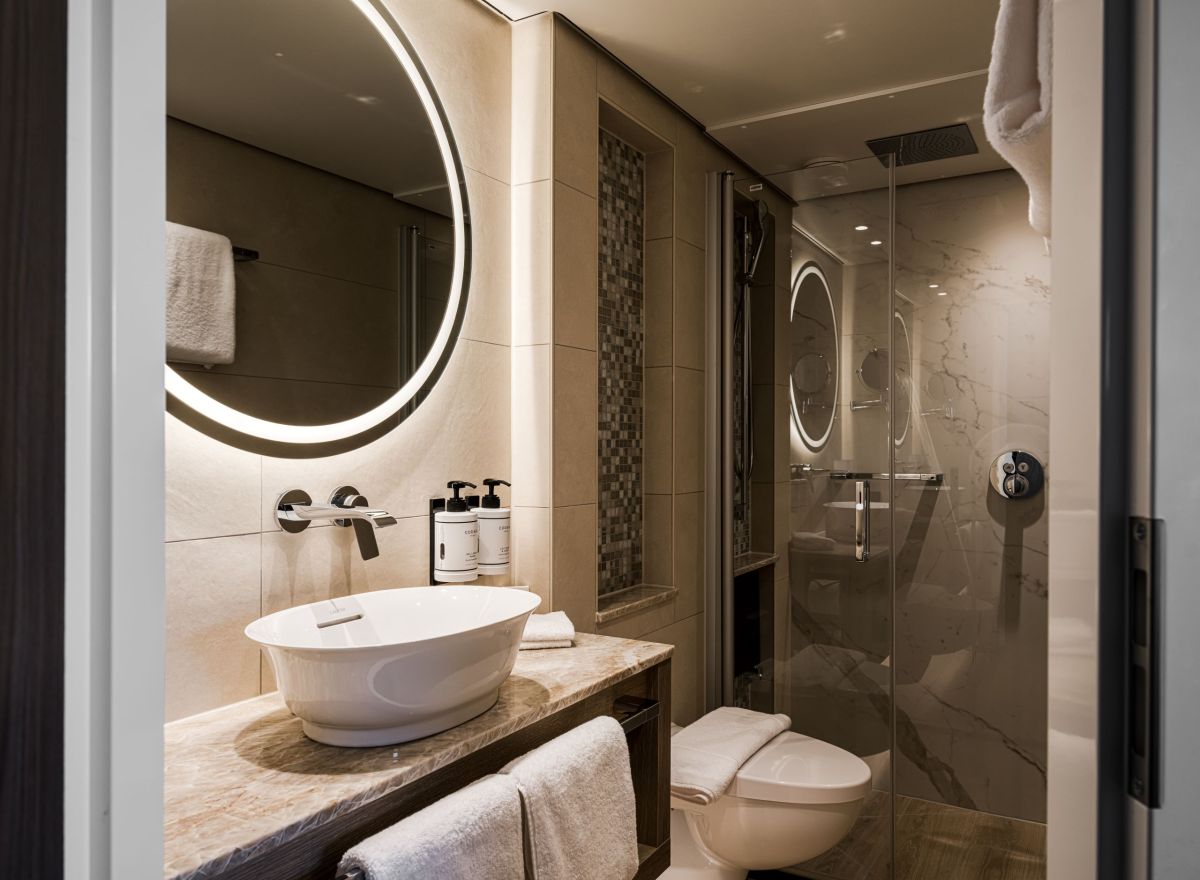 Luxury cruise bathroom with a round backlit mirror, vessel sink, marble-style surfaces, and a glass-enclosed rainfall shower.