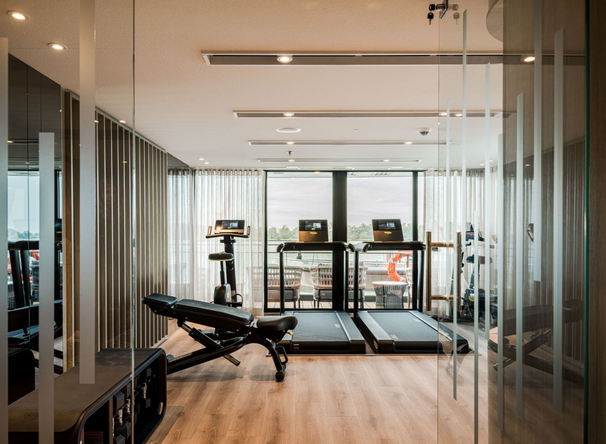 Modern onboard gym with treadmills, a stationary bike, free weights, and floor-to-ceiling windows overlooking the river.