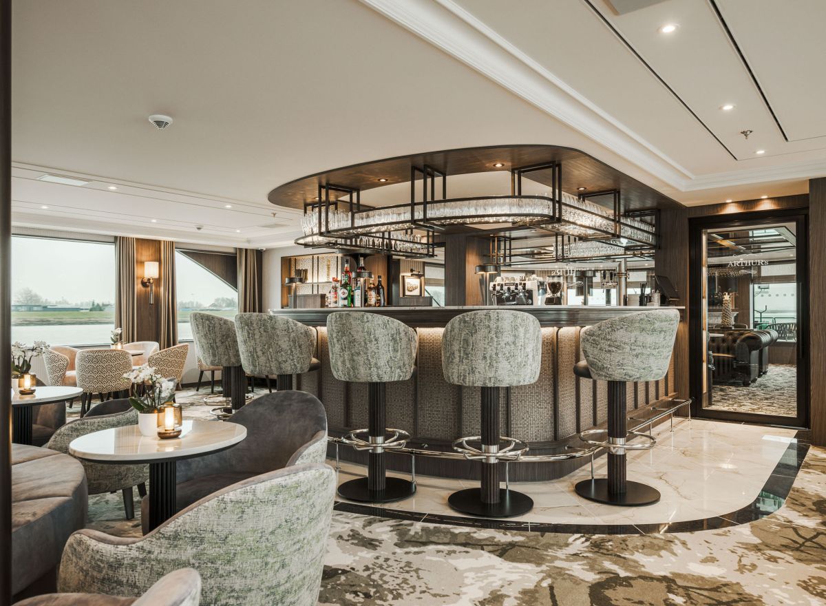 Modern onboard bar and lounge with plush seating, patterned barstools, mirrored ceiling details, and large windows with river views.