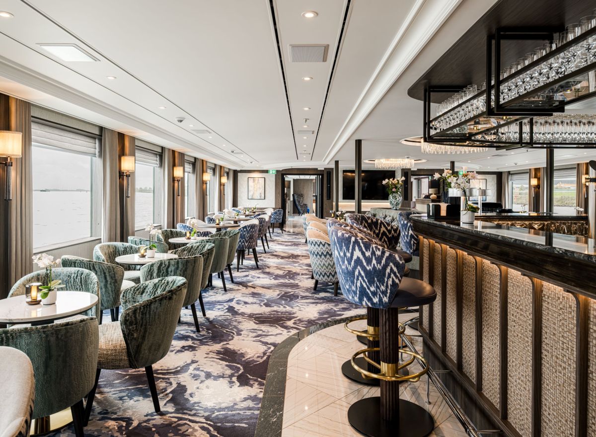 Modern onboard bar and lounge with plush seating, patterned barstools, mirrored ceiling details, and large windows with river views.
