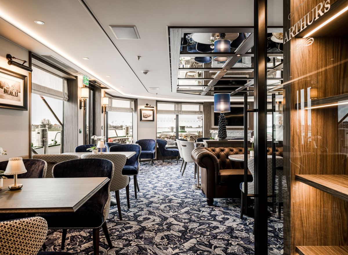 Refined onboard dining lounge with blue velvet chairs, patterned upholstery, and large riverside windows.